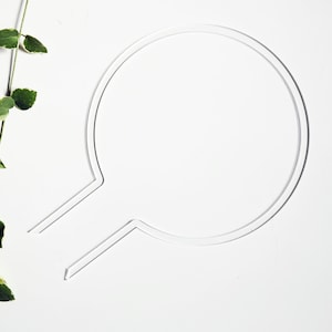 以下が含まれることがあります： 白い背景に、薄い銀色の円形の輪郭が1つと、その円の下から伸びる薄い銀色の線が2本あります。画像の左側には、白い斑点のある緑色のつるがあります。