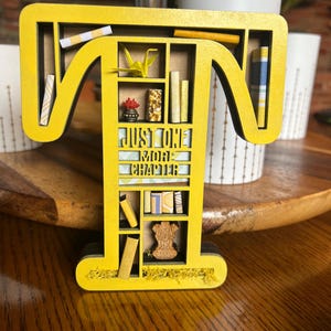 May include: A yellow wooden letter "T" shaped bookshelf with miniature books, a paper crane, and the text "JUST ONE MORE CHAPTER". The bookshelf is decorated with small objects and is on a wooden surface.
