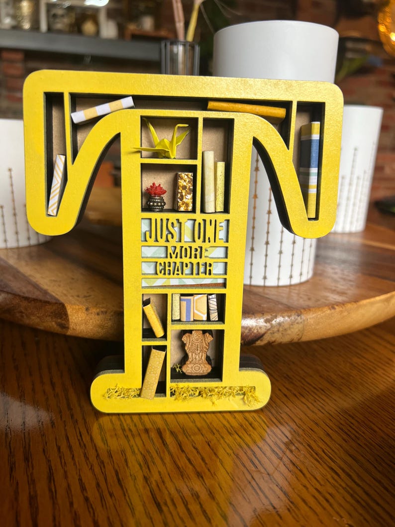 May include: A yellow wooden letter "T" shaped bookshelf with miniature books, a paper crane, and the text "JUST ONE MORE CHAPTER". The bookshelf is decorated with small objects and is on a wooden surface.