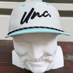 May include: A light gray baseball cap featuring the black embroidered text "Unc." on the front panel. The cap includes a light blue brim, a black and white braided rope detail across the base of the crown, and light blue eyelets on top.