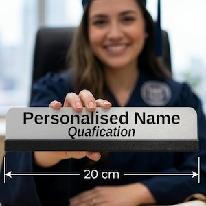 May include: A graduation gift with a personalised name and qualification on a rectangular plaque, measuring 20 cm. The plaque is held up by a person wearing a graduation gown and cap. A rolled diploma rests on the desk.
