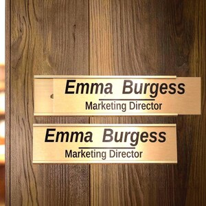 May include: Two gold-coloured nameplates on a wooden door, each bearing the name "Emma Burgess" and the title "Marketing Director" in black lettering. The door has a visible wood grain pattern.