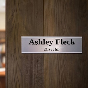 May include: A silver nameplate on a wooden door reads "Ashley Fleck, Director" in black text. The door is made of dark brown wood with vertical grain patterns. The door is slightly ajar, revealing a glimpse of an office interior.