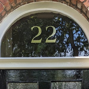 May include: A house entrance with a black door and a semi-circular window above. The window displays the number "22" in gold. The window is framed by a white arch, set against a red brick wall. The door has a glass panel.