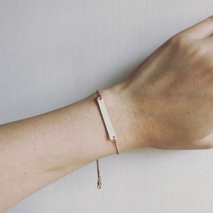 May include: A rose gold bar bracelet is displayed on a wrist. The bracelet has a rectangular bar pendant and a fine chain. The adjustable chain ensures a comfortable fit. This bracelet is a simple and elegant piece of jewellery.