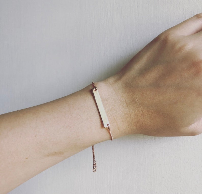 May include: A rose gold bar bracelet is displayed on a wrist. The bracelet has a rectangular bar pendant and a fine chain. The adjustable chain ensures a comfortable fit. This bracelet is a simple and elegant piece of jewellery.