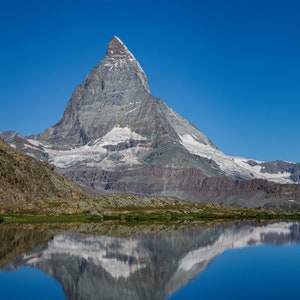 May include: A towering mountain with a snow-covered peak is mirrored in a tranquil, dark blue lake. The mountain's grey and brown slopes contrast with the clear, bright blue sky. The image captures a serene landscape.