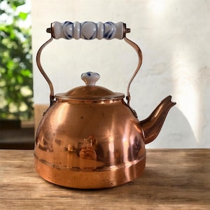 Puede incluir: Una tetera de cobre con un asa y un pomo de cerámica blanca y azul. La tetera tiene un cuerpo redondeado, un pico curvo y un asa que se arquea hacia arriba. La superficie de cobre refleja el entorno.
