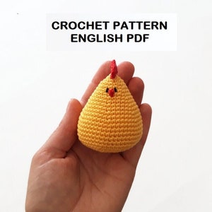 Könnte beinhalten: Ein gehäkeltes gelbes Küken mit rotem Kamm und schwarzen Augen. Das Küken wird in einer Hand gehalten. Häkelanleitung Englisch PDF.