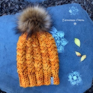 May include: A bright orange and yellow knitted beanie with a brown and white faux fur pom pom. The beanie has a small white tag with the word "Evergreen" on it. The beanie is sitting on a blue surface with white designs.