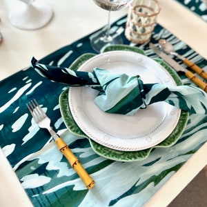 May include: A table setting with a green and white leaf-patterned runner, a white plate with a green scalloped plate underneath, and a matching napkin. Silverware with bamboo-style handles is placed on the table.