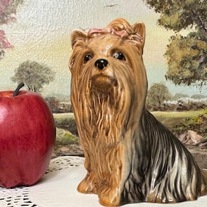 May include: A ceramic Yorkshire Terrier figurine with a pink bow on its head sits next to a red apple. The dog has brown and black fur and is posed in a sitting position. The background is a painting of a landscape.