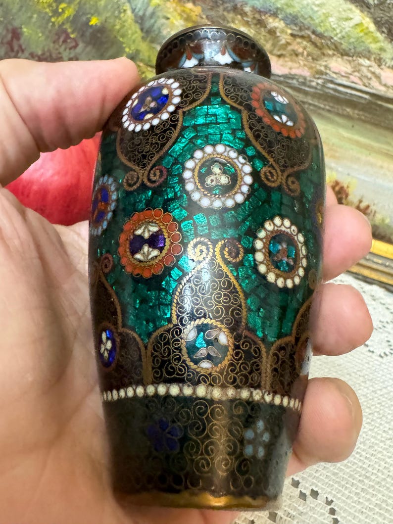 May include: A small, decorative vase with a green, black, and gold cloisonn&eacute; design. The vase is hand-painted with intricate details and features a mosaic-like pattern.