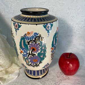 May include: A white ceramic vase with a blue, green, and pink floral pattern. The vase has a black rim and base and is decorated with gold accents.