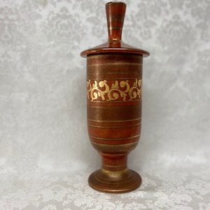 May include: A brown ceramic jar with a gold lid and gold floral design. The jar has a round base and a tall, slender body.