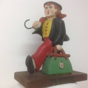 May include: A wooden figurine of a person wearing a black coat, red pants, and a yellow vest. The person is holding a green suitcase and a black hook. The figurine is standing on a wooden base.