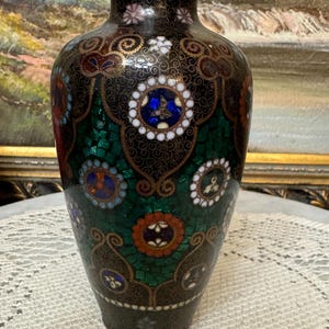 May include: A black cloisonn&eacute; vase with intricate green, blue, red, and white floral patterns. The vase has a round body and a narrow neck.