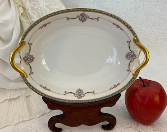 May include: An oval serving bowl with gold handles and a floral design in pink, blue, and gold. The bowl is on a wooden stand.