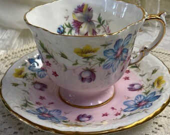 May include: A delicate teacup and saucer set with a pink and white floral pattern. The cup and saucer have a gold trim and feature a variety of flowers, including blue, yellow, and pink.