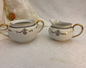 May include: A white porcelain sugar bowl and creamer set with gold trim and floral designs. The set features a delicate floral pattern with pink and blue accents.