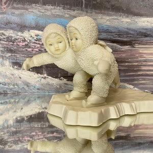 May include: Two white porcelain figurines of children wearing winter clothing and ice skates, standing on a white base that resembles a frozen pond. The figurines are facing each other, with the child on the left looking to the right and the child on the right looking to the left.