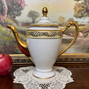 May include: A white porcelain teapot with gold trim and a floral design. The teapot has a gold handle and spout. It is sitting on a white lace doily.