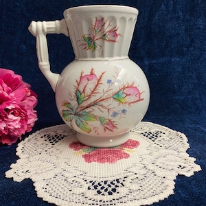 May include: White ceramic pitcher with a floral design featuring pink, green, and blue flowers. The pitcher has a round body and a flared top with a handle.