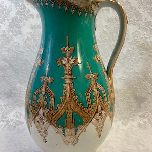May include: A large, green and gold ceramic pitcher with a decorative design of arches and spires. The pitcher has a wide mouth and a handle.