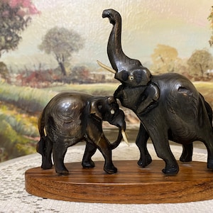 May include: A wooden sculpture of two elephants, one larger than the other, standing on a wooden base. The elephants are carved in a dark brown color and have ivory tusks.