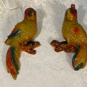 May include: Two yellow, red, and green ceramic parrot figurines perched on branches. The parrots have black eyes and are facing different directions.