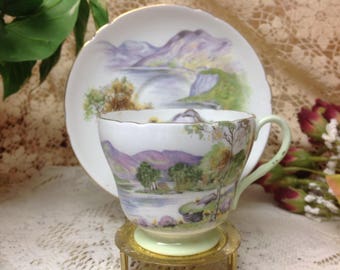 May include: A vintage teacup and saucer set with a delicate floral design featuring a lake and mountains. The cup and saucer are white with a light green rim and a gold trim.