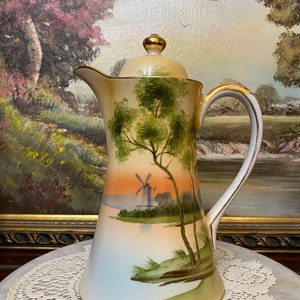 May include: A white porcelain coffee pot with a gold trim and a hand-painted scene of a windmill and trees. The pot has a lid with a gold knob.