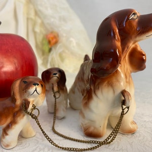 May include: Three ceramic dog figurines with brown and white fur. The largest dog is standing with its head turned to the side. The two smaller dogs are sitting and have gold chains attached to their collars.