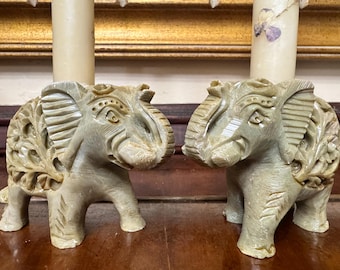 May include: Two carved stone elephant candle holders. The elephants are light green with detailed carvings on their ears and bodies. Each elephant has a candle in its back. The candle holders are on a wooden surface.
