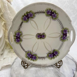 May include: A round white porcelain serving plate with two side handles, featuring a central cluster and six outer clusters of purple violet flowers with green leaves, connected by thin gold lines and a decorative silver filigree border.