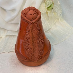 May include: A terracotta figurine of a person wearing a hooded cloak. The figurine is hand-crafted and has a detailed face and textured clothing.