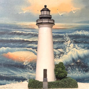 May include: A white lighthouse figurine with a black top and a small green bush at its base. The lighthouse is set against a backdrop of a painted ocean scene with waves and seagulls.