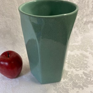 May include: A tall, light green ceramic vase with a geometric, faceted design. The vase has a speckled finish and a slightly rounded top.