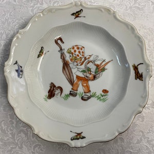 May include: A white porcelain plate with a scalloped edge features a colorful illustration of a girl carrying a basket of flowers and a walking stick. The plate is decorated with butterflies, a squirrel, and a fish.