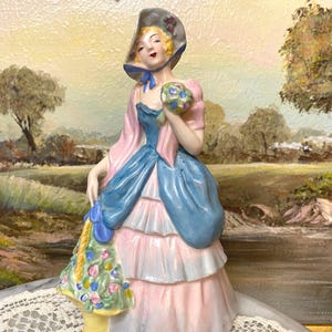 May include: A porcelain figurine of a woman in a pink and blue dress, holding a bouquet of flowers. She is wearing a blue hat with a white brim and a blue ribbon. The figurine is standing on a white surface with a lace pattern.