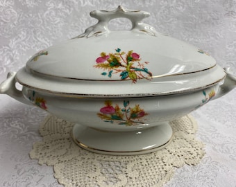 May include: White porcelain serving bowl with a lid and floral design. The bowl has a gold trim around the edge and a floral pattern with pink, green, and yellow flowers.