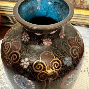 May include: A small, dark brown vase with a blue and white enamel design. The vase is decorated with gold swirls and floral patterns.