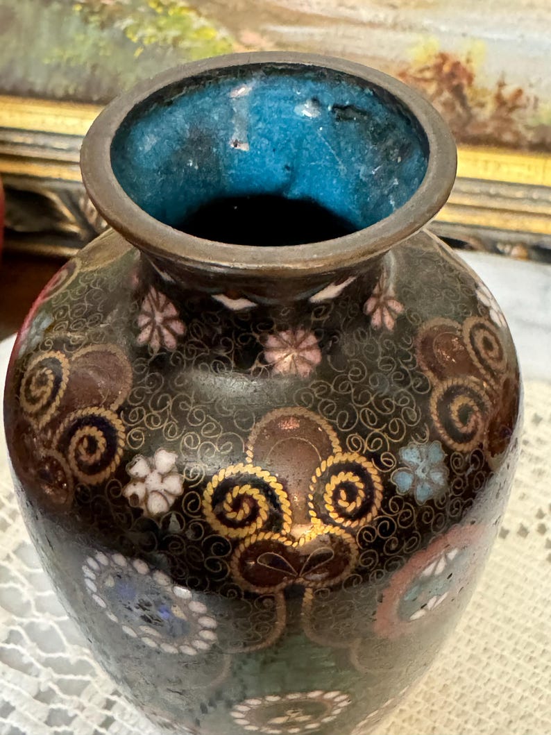 May include: A small, dark brown vase with a blue and white enamel design. The vase is decorated with gold swirls and floral patterns.