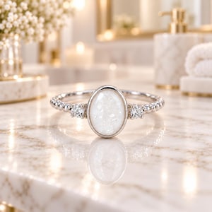 Puede incluir: Un anillo de tono plateado con una piedra druzy blanca central de forma ovalada en un engaste liso. La banda tiene una textura de cuentas y está acentuada a cada lado por una pequeña piedra redonda transparente facetada en engaste de garras.
