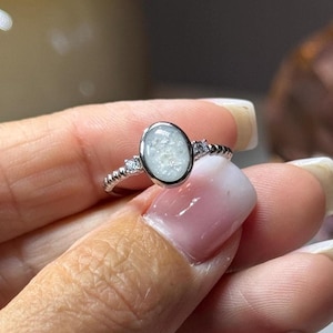 May include: A silver-toned ring featuring an oval-shaped center stone with a textured, pearlescent white appearance. The band has a beaded design and is accented by two small, clear, round stones positioned on either side of the central setting.