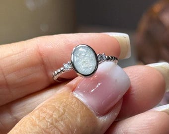 Könnte beinhalten: Ein silberfarbener Ring mit einem zentralen, ovalen Stein mit texturierter, schimmernder weißer Oberfläche. Die Ringschiene hat ein Perlendesign und wird von zwei kleinen, klaren, rund geschliffenen Steinen flankiert.
