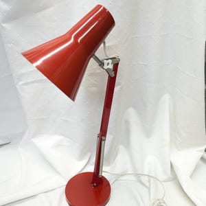 K&ouml;nnte beinhalten: Eine rote Schreibtischlampe mit konischem Schirm und rundem Fu&szlig;. Die Lampe hat einen flexiblen Arm und ein wei&szlig;es Netzkabel. Die Lampe ist aus Metall und hat eine gl&auml;nzende Oberfl&auml;che. Die Lampe ist f&uuml;r den Schreibtischgebrauch konzipiert.