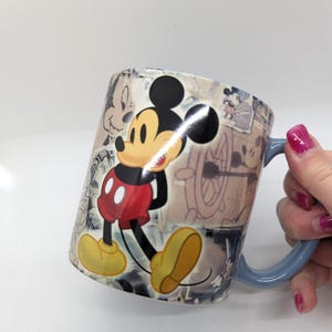 Op de afbeelding: Een keramische koffiemok met een klassiek Mickey Mouse-ontwerp. De mok heeft een lichtblauw handvat en is versierd met afbeeldingen van Mickey Mouse in verschillende poses en vintage schetsen. De primaire kleuren zijn rood, geel, zwart en wit.