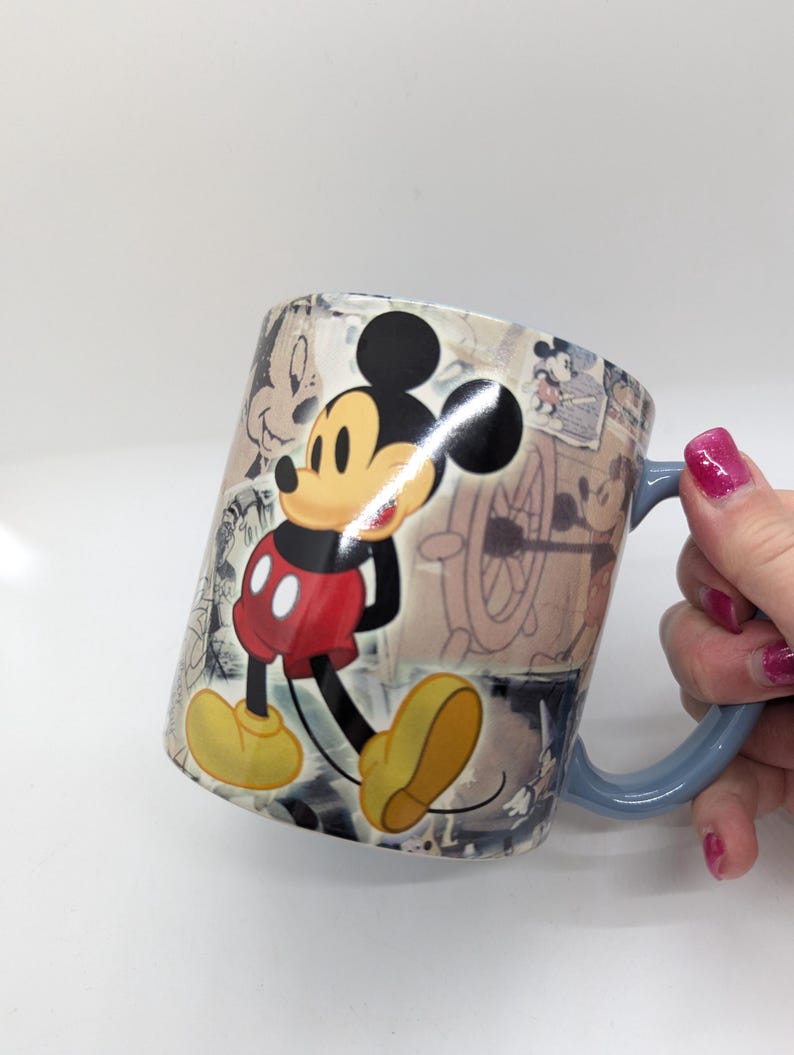 Op de afbeelding: Een keramische koffiemok met een klassiek Mickey Mouse-ontwerp. De mok heeft een lichtblauw handvat en is versierd met afbeeldingen van Mickey Mouse in verschillende poses en vintage schetsen. De primaire kleuren zijn rood, geel, zwart en wit.