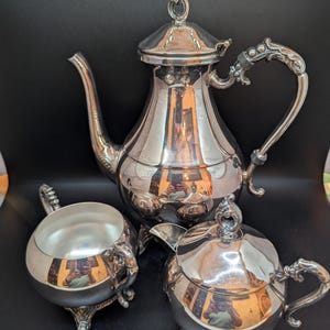 Op de afbeelding: Een zilveren theeservies met sierlijke details. De set bevat een theepot, een suikerpot en een melkkan. Elk stuk heeft decoratieve handvatten en staat op kleine voetjes. De reflecterende oppervlakken creëren een heldere, elegante uitstraling.